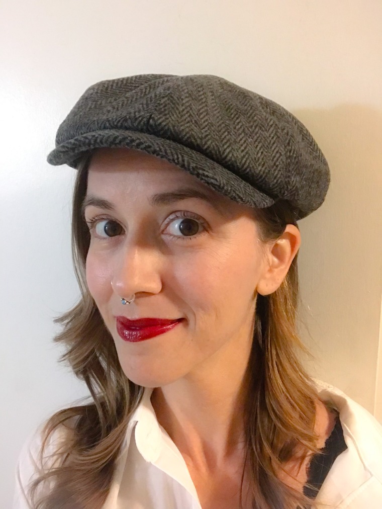 A photo of Sarah Maaske, a white nonbinary femme with light brown hair and brown eyes. They are wearing a white shirt with buttons and a grey cap. They have red lipstick on.